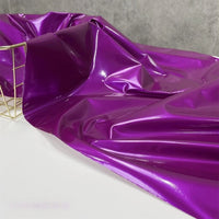 A yard of Longan Craft's Glossy Stretch Vinyl Fabric in purple is draped over a white surface, with a gold wire basket on the left and a gray textured background behind. This shiny PU vinyl is perfect for making jackets.
