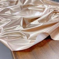 A sheet of Longan Craft Glossy Stretch Vinyl Fabric By The Yard in metallic gold, a shiny PU material ideal for jackets, is displayed with wrinkles on a wooden surface.
