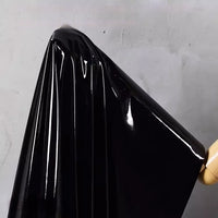 A yard of Longan Craft’s Glossy Stretch Vinyl Fabric in shiny black, with a mirror-like finish and sleek folds, hangs on a wooden hanger against a light gray wall—ideal for jackets.