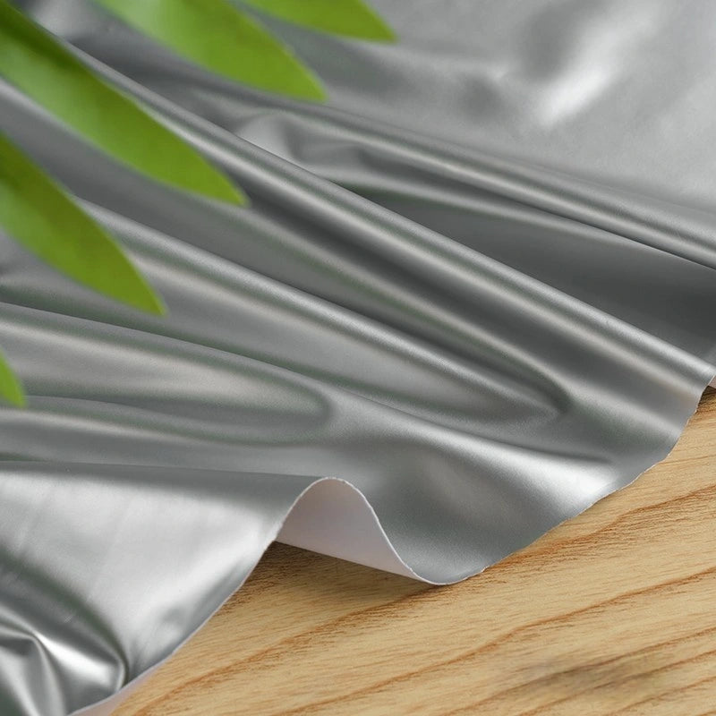 Longan Craft's Glossy Stretch Vinyl Fabric by the Yard—solid color shiny PU vinyl, perfect for jackets—rests on a wooden surface, partially covered by green leaves in the upper left corner.
