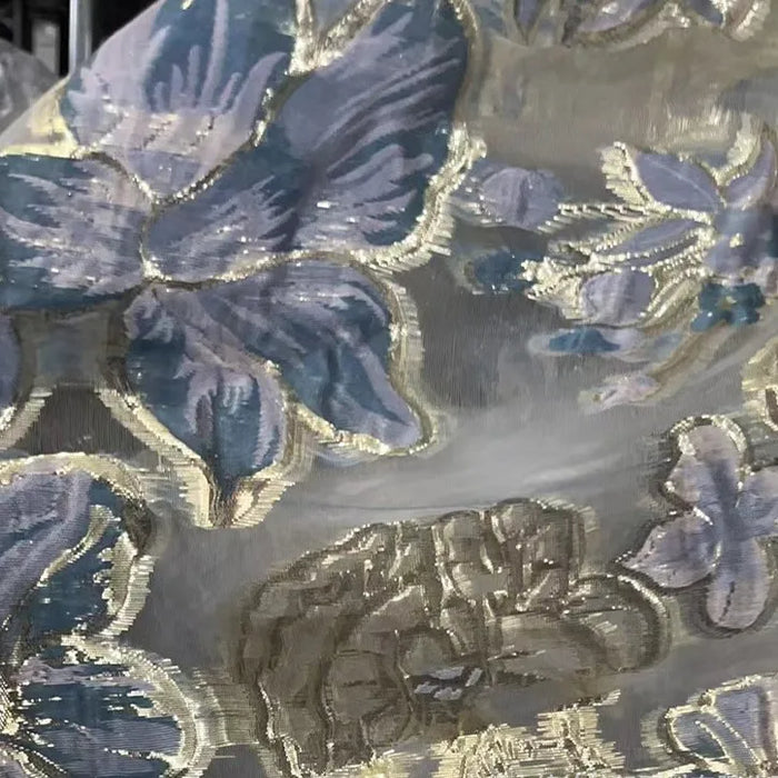 Golden Floral Jacquard Organza Fabric by Longan Craft features large purple, blue, and beige flowers with gold accents on a sheer organza. Perfect for dressmaking, this material is sold by the yard and showcases an elegant floral pattern.