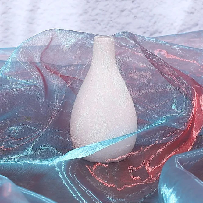 A matte white teardrop-shaped vase stands on Longan Craft's Gradient Organza Fabric By The Yard, a lightweight ombre organza cloth in blue and pink tones, set against a textured light background.