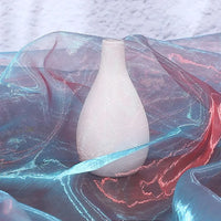 A matte white teardrop-shaped vase stands on Longan Craft's Gradient Organza Fabric By The Yard, a lightweight ombre organza cloth in blue and pink tones, set against a textured light background.