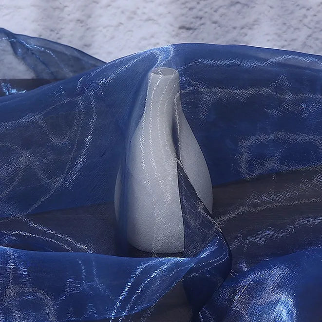 A white ceramic vase, partially wrapped in Longan Craft’s Gradient Organza Fabric By The Yard, sits on a surface with a textured white background and hints of the soft ombre organza cloth material shimmering gently.