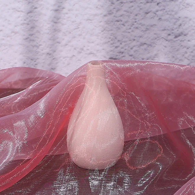 A pale pink ceramic vase rests on a surface draped with Longan Craft’s Gradient Organza Fabric By The Yard, a lightweight ombre organza cloth, with a textured light purple wall in the background.