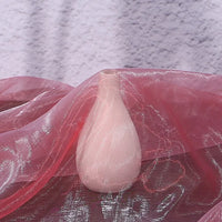 A pale pink ceramic vase rests on a surface draped with Longan Craft’s Gradient Organza Fabric By The Yard, a lightweight ombre organza cloth, with a textured light purple wall in the background.