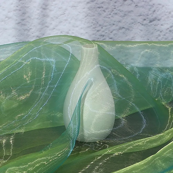 A white ceramic vase stands on a surface, partially covered and surrounded by Longan Craft's Gradient Organza Fabric By The Yard in green and blue ombre tones, featuring a wavy, reflective texture. A light gray wall is in the background.