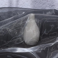 A white ceramic vase is draped with Longan Craft’s Gradient Organza Fabric, a lightweight ombre organza cloth, shimmering in black and silver patterns against a textured, light-colored background.