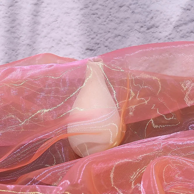 A teardrop-shaped candle is partly wrapped in Longan Craft’s Gradient Organza Fabric By The Yard, a soft, lightweight ombre organza cloth, set against a textured pale background.