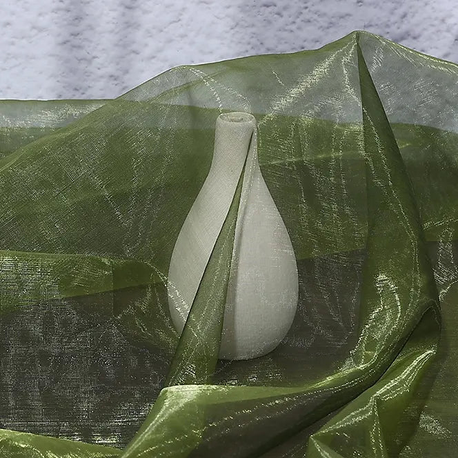 A white ceramic vase is elegantly draped with Longan Craft's Gradient Organza Fabric By The Yard, a lightweight ombre organza cloth, its sheer green material set against a textured light background.