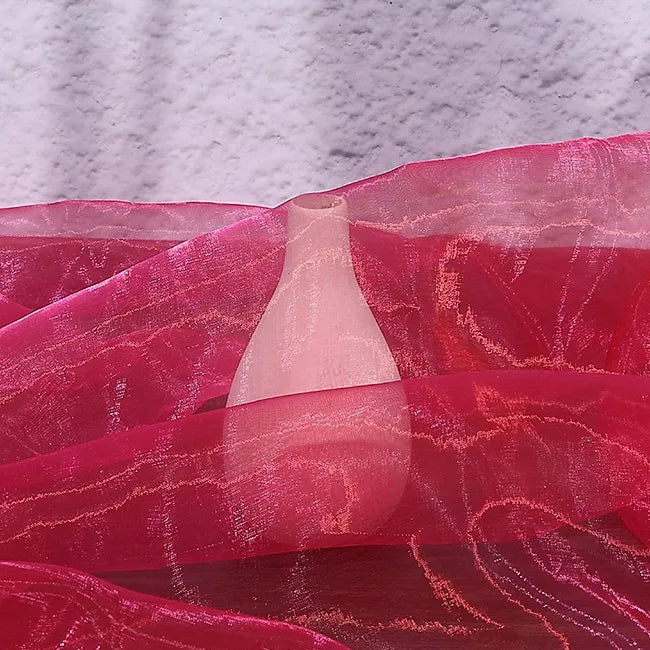 A white ceramic vase is gently draped with Longan Craft’s Gradient Organza Fabric By The Yard, a lightweight ombre organza material, set against a textured light purple wall with vibrant pink cloth cascading around the surface.