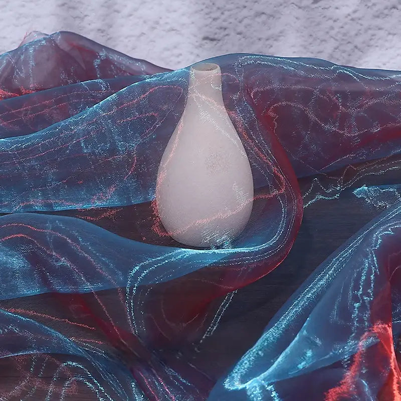 A white ceramic vase rests on a dark surface, partly draped with Longan Craft's Gradient Organza Fabric By The Yard—lightweight ombre organza cloth in blue and red hues—its soft sheen reminiscent of bridal elegance against a textured background.