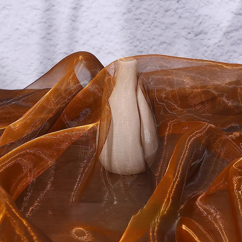A small beige ceramic vase rests on a surface draped with Longan Craft’s Gradient Organza Fabric By The Yard, its lightweight ombre material in orange-brown hues contrasting with a textured white wall in the background.