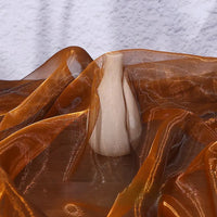 A small beige ceramic vase rests on a surface draped with Longan Craft’s Gradient Organza Fabric By The Yard, its lightweight ombre material in orange-brown hues contrasting with a textured white wall in the background.