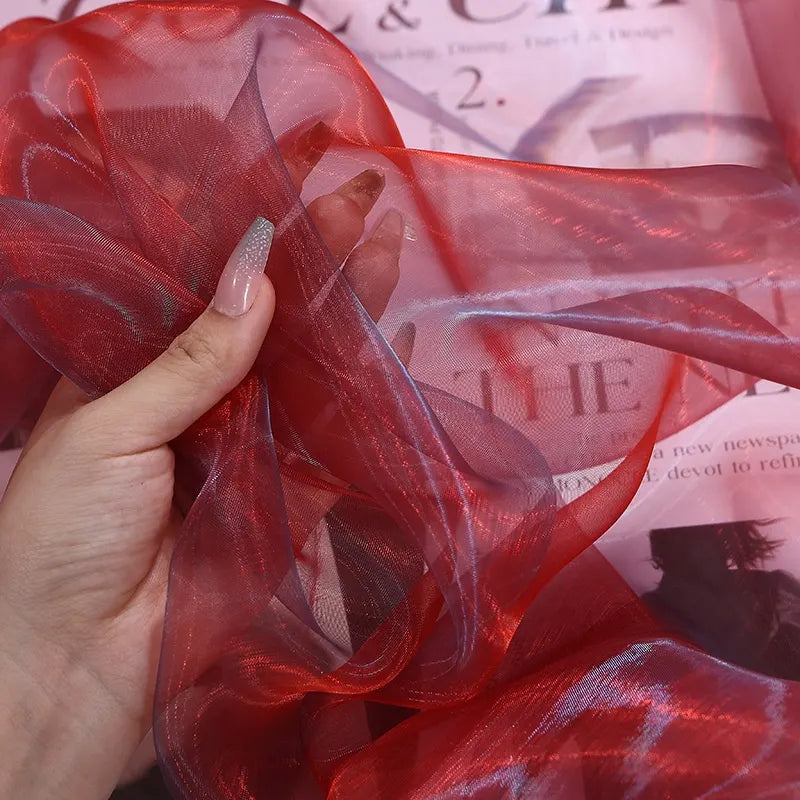 A hand with long, manicured nails holds Longan Craft’s Gradient Organza Fabric By The Yard, a lightweight ombre organza cloth, with a pink newspaper or magazine in the background.