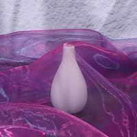 A white, tulip-shaped vase is partially draped in Longan Craft's Gradient Organza Fabric By The Yard, a soft ombre organza cloth, creating delicate folds and reflections against a textured white background.