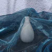 A small, white, smooth vase sits on Longan Craft’s Gradient Organza Fabric By The Yard—a lightweight, ombre organza with a soft, sheer blue surface—against a textured light gray backdrop.