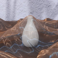 A white matte ceramic vase with a narrow neck rests on Longan Craft's Gradient Organza Fabric By The Yard, a lightweight ombre organza cloth featuring blue highlights and soft folds, against a textured light gray background.