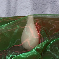 A small, white, matte vase sits on Longan Craft’s Gradient Organza Fabric By The Yard, featuring a shiny green and red ombre design, with a textured gray background behind it.