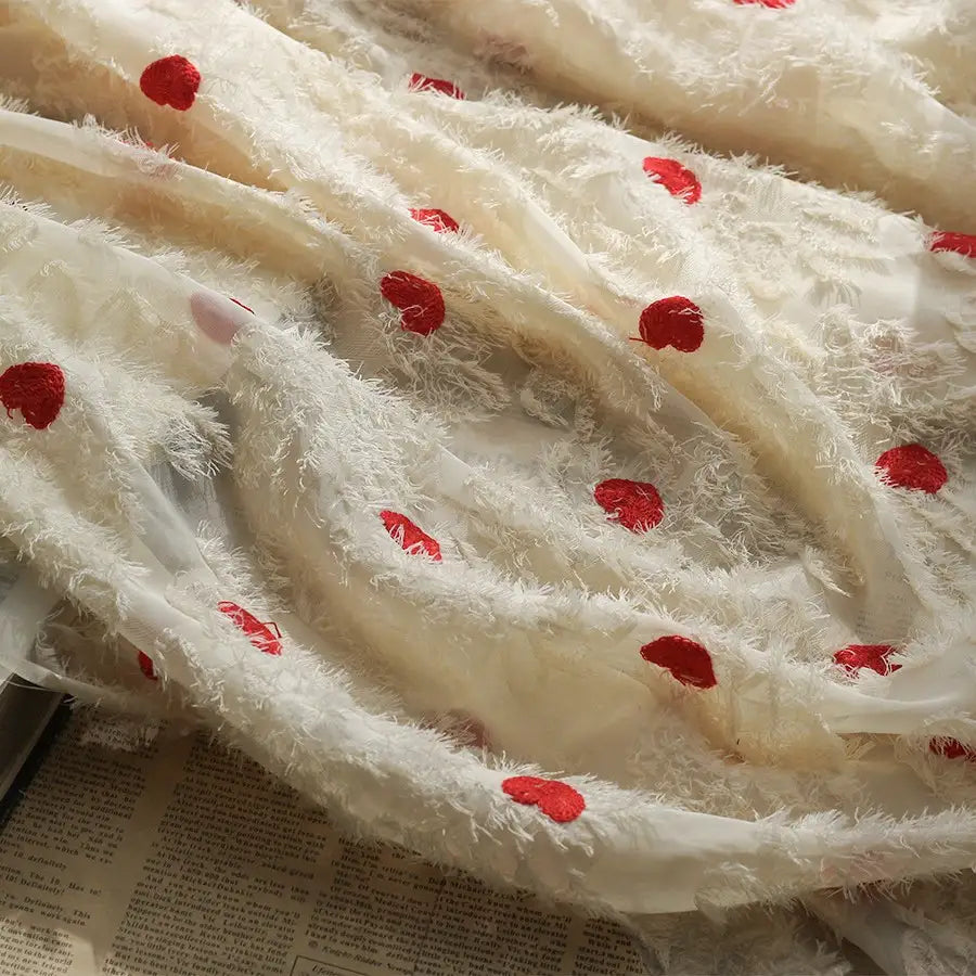 A close-up of Longan craft’s Heart Embroidered Tassel Chiffon Fabric by the yard, a sheer jacquard fabric with fluffy details and red dots, draped over an open newspaper—ideal sewing material for spring and summer women’s dresses.