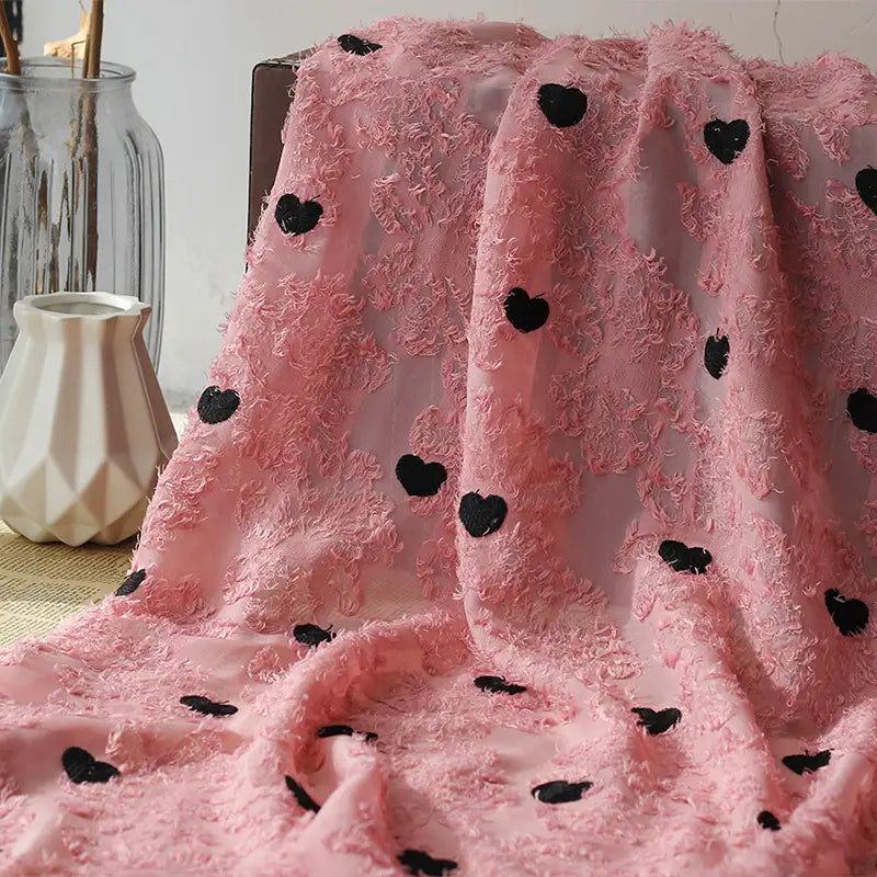 A Heart Embroidered Tassel Chiffon Fabric by Longan craft, featuring black heart patterns, is draped over a chair beside geometric white and clear glass vases. Ideal jacquard sewing material for spring and summer women's dresses.