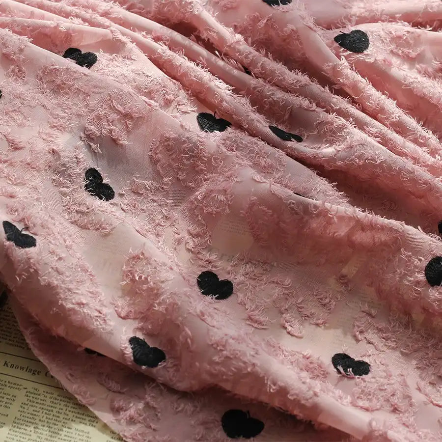 A close-up of Longan craft's Heart Embroidered Tassel Chiffon Fabric by the yard, featuring textured fuzzy hearts and black heart patterns, draped over a surface with faint text beneath—ideal Jacquard fabric for spring and summer dresses.