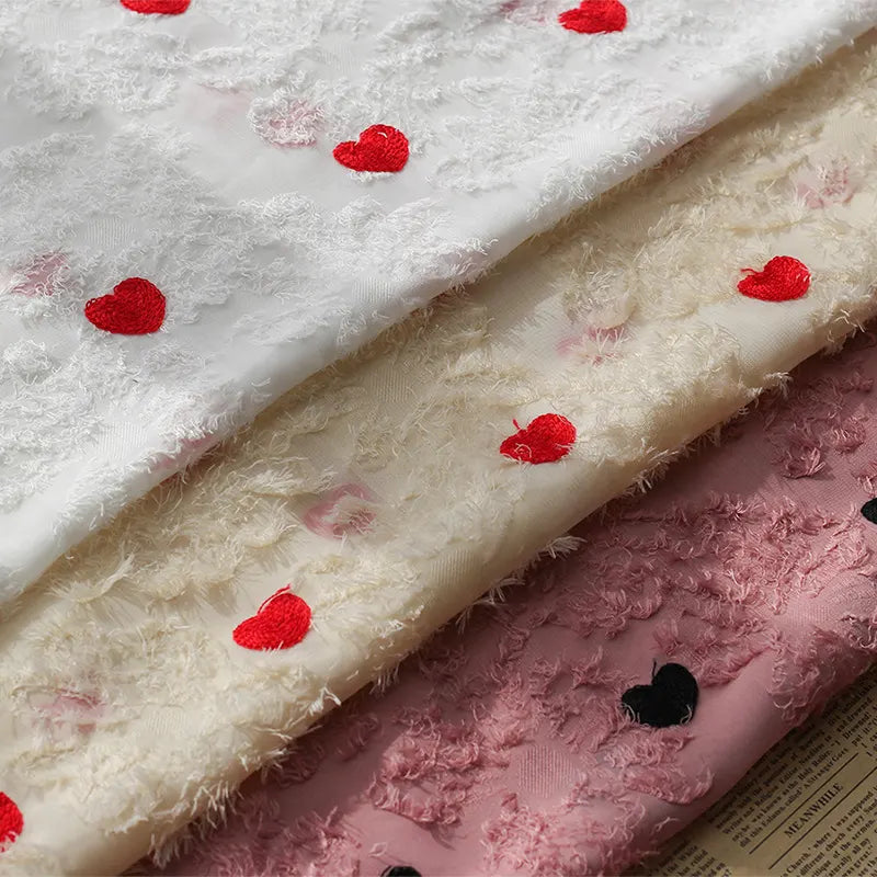 Three layers of Longan craft Heart Embroidered Tassel Chiffon Fabric by the yard—white, cream, and pink with red and black heart embroidery—are stacked on a printed surface; ideal for spring/summer women’s dress sewing.