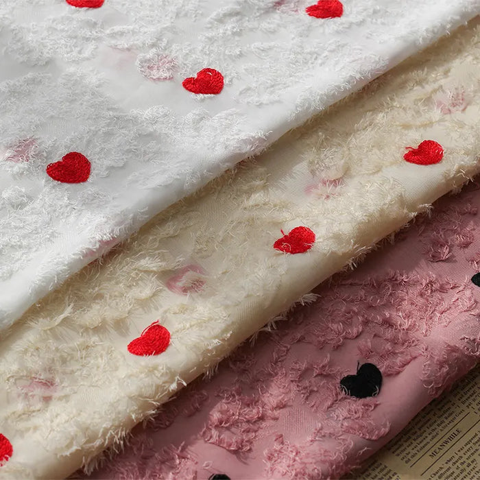 Three layers of Longan craft Heart Embroidered Tassel Chiffon Fabric by the yard—white, cream, and pink with red and black heart embroidery—are stacked on a printed surface; ideal for spring/summer women’s dress sewing.