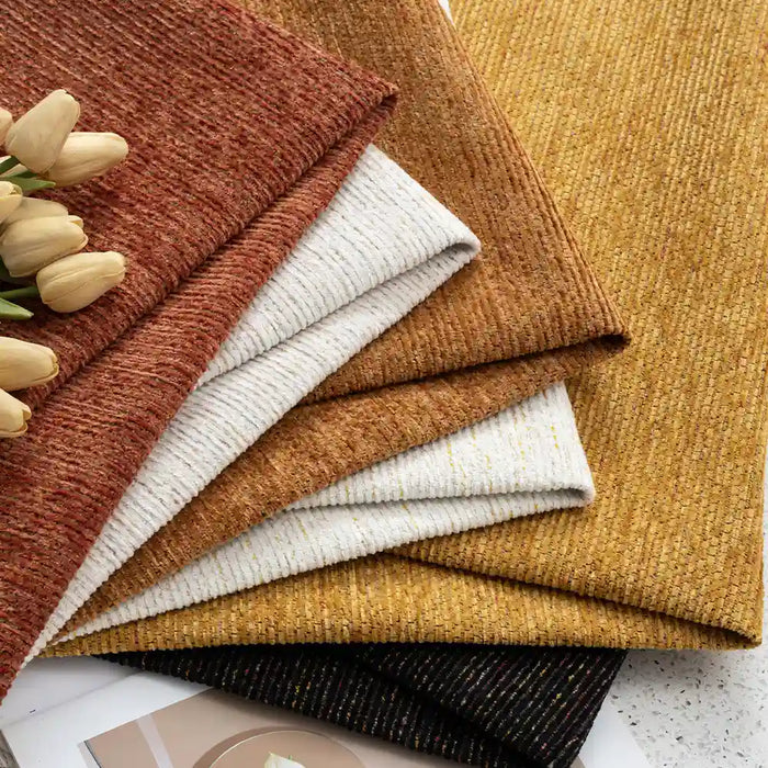 A neatly arranged stack of Longan Craft Heavy Weight Slub Chenille Fabric in rust, beige, mustard, gold, white, and black sits by pale flowers on a light background—perfect premium upholstery material sold by the yard.