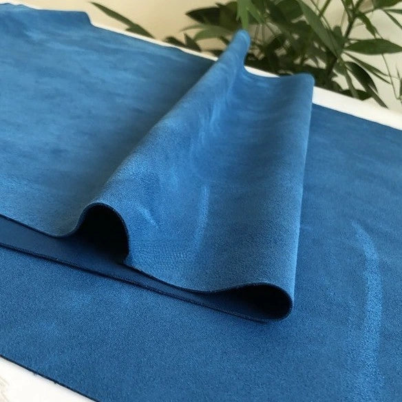 A sheet of Longan Craft Heavyweight Microfiber Faux Suede Fabric By The Yard in blue is draped and folded on a white surface, with green leafy plants softly blurred in the background.