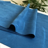 A sheet of Longan Craft Heavyweight Microfiber Faux Suede Fabric By The Yard in blue is draped and folded on a white surface, with green leafy plants softly blurred in the background.