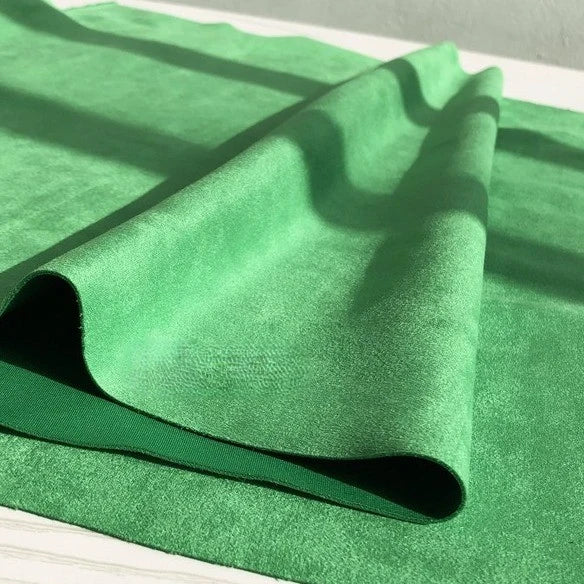 A close-up of a folded piece of Longan Craft Heavyweight Microfiber Faux Suede Fabric By The Yard in bright green, resting on a matching green surface, with sunlight softly casting shadows across the multicolor brushed microsuede material.