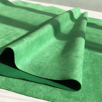 A close-up of a folded piece of Longan Craft Heavyweight Microfiber Faux Suede Fabric By The Yard in bright green, resting on a matching green surface, with sunlight softly casting shadows across the multicolor brushed microsuede material.