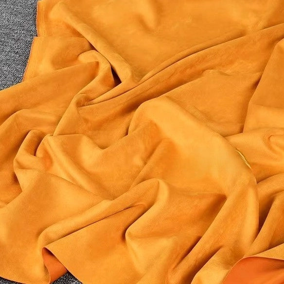 The Longan Craft Heavyweight Microfiber Faux Suede Fabric By The Yard, seen here in a soft orange shade, is draped and spread out on a gray surface, displaying gentle folds and wrinkles in its multicolor brushed microsuede material—perfect for DIY clothing projects.