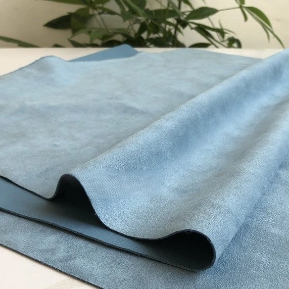 A close-up of Longan Craft’s Heavyweight Microfiber Faux Suede Fabric By The Yard in light blue, featuring a soft, suede-like brushed surface and heavyweight texture, is partially folded on a white surface. Soft green plant leaves appear blurred in the background.