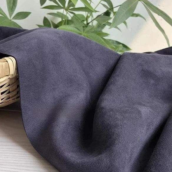 A piece of Longan Craft Heavyweight Microfiber Faux Suede Fabric By The Yard in dark gray is draped over the edge of a wicker basket, with green leafy plants softly blurred in the background.