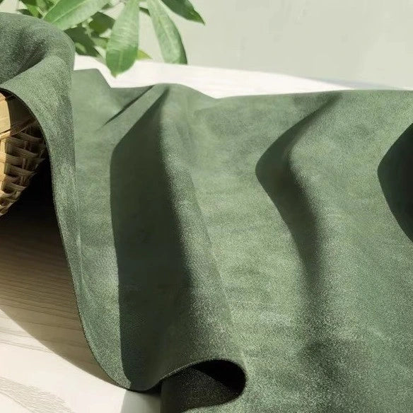 A piece of green Heavyweight Microfiber Faux Suede Fabric By The Yard from Longan Craft is draped across a light wooden surface, accompanied by part of a woven basket on the left and green leaves in the background.
