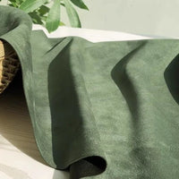 A piece of green Heavyweight Microfiber Faux Suede Fabric By The Yard from Longan Craft is draped across a light wooden surface, accompanied by part of a woven basket on the left and green leaves in the background.