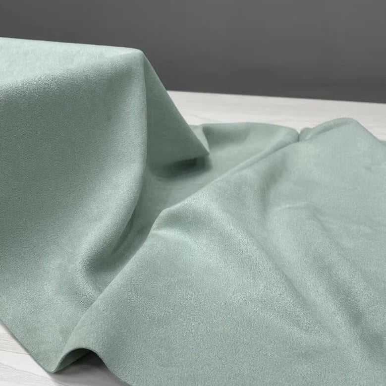 A yard of Longan Craft’s Heavyweight Microfiber Faux Suede Fabric in light green is artfully draped over a white surface, forming gentle folds and shadows against a plain grey background.