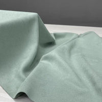 A yard of Longan Craft’s Heavyweight Microfiber Faux Suede Fabric in light green is artfully draped over a white surface, forming gentle folds and shadows against a plain grey background.