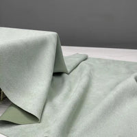 A heavyweight microfiber faux suede fabric by Longan Craft, available by the yard in multicolor brushed microsuede, is displayed in light green draped over a light gray surface. Gentle folds reveal its soft, imitation suede texture against a plain gray background. This luxurious yet durable Longan Craft fabric is an excellent choice for any décor or DIY clothing project.