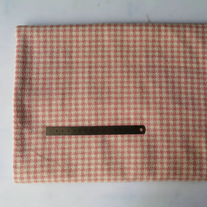 A piece of Longan Craft Houndstooth Mammoth Flannel Fabric in pink and cream rests folded on a white surface, with a metal ruler on top for scale. This brushed polyester cotton flannelette is sold by the yard, perfect for cozy fall projects.