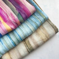 Three pieces of Longan Craft Irregular Stripe Printed Ramie Fabric By The Yard, featuring lightweight and breathable natural ramie material with watercolor-style stripes in pink, blue, and earthy tones, are neatly stacked on a light surface. Each artistic piece showcases the unique irregular striped design, making them ideal for dressmaking.