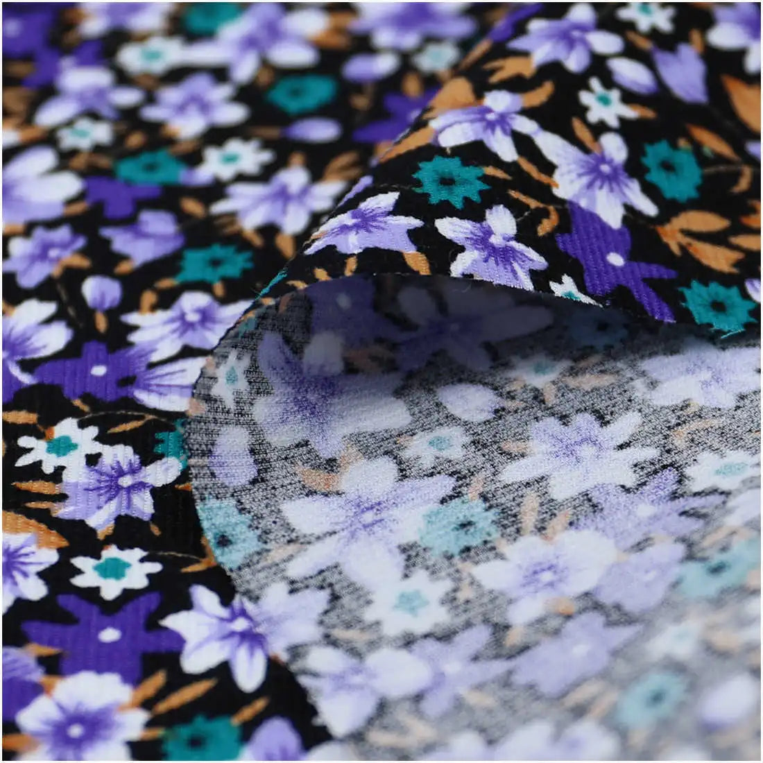 Close-up of Lily Printed Cotton Corduroy Fabric by Longan Craft, featuring purple, aqua, white, and tan floral designs on black. Part of this fine wale corduroy is flipped to show the faded reverse side. Ideal for tops.