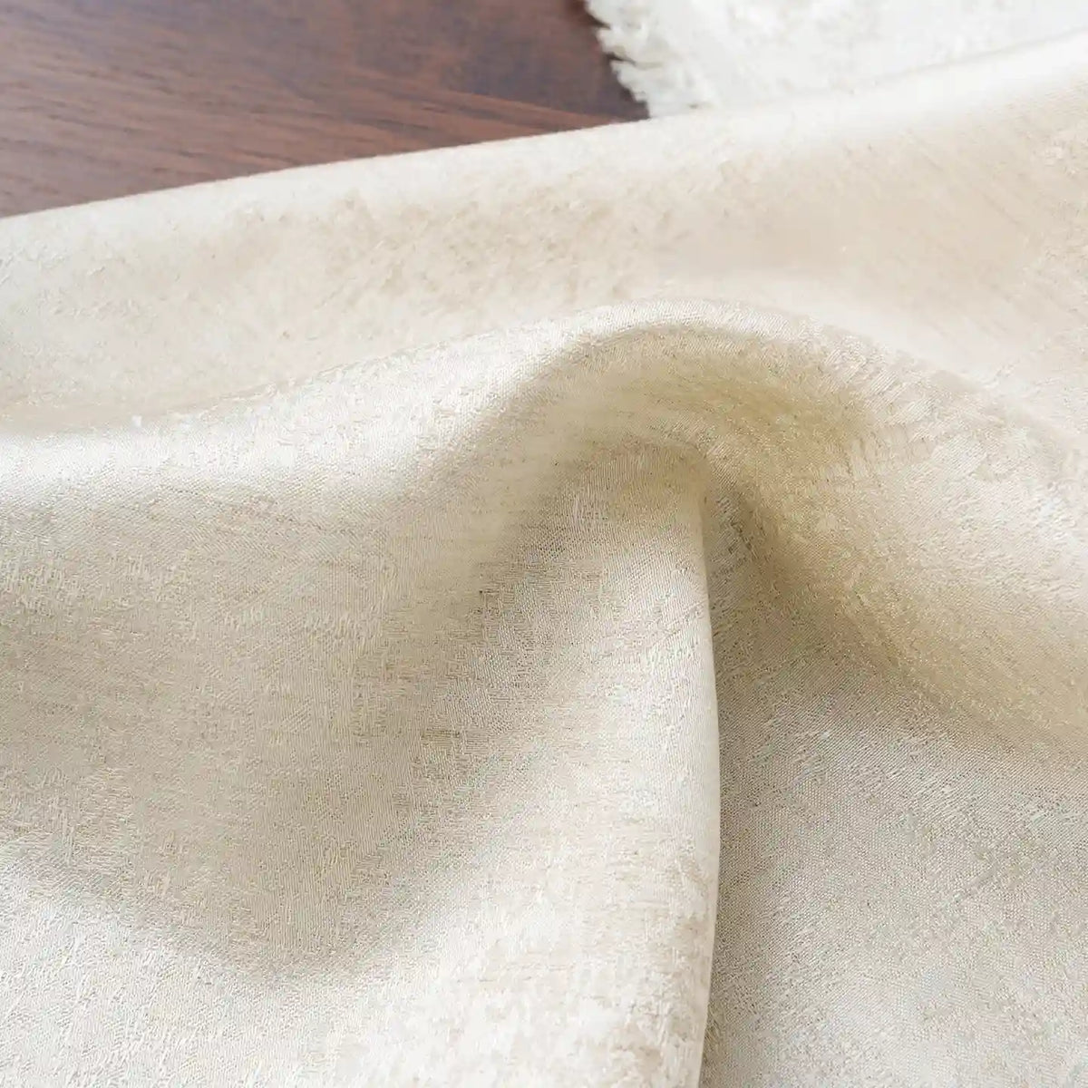 A close-up of Longan Craft’s Linen Viscose Jacquard Satin Fabric by the yard, featuring a subtle floral pattern in cream tones, softly draped on a dark wooden surface and partially overlapping a fringed light cloth—an ideal luxury satin fabric for elegant dressmaking and refined sewing projects.