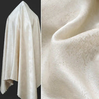 A cream-colored Longan Craft Linen Viscose Jacquard Satin Fabric By The Yard is elegantly draped over a stand on the left, with a close-up on the right highlighting its subtle floral pattern and luxurious sheen. This rayon satin material is perfect for creating sophisticated dresses and other refined garments.