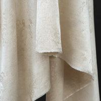 A close-up view of the Linen Viscose Jacquard Satin Fabric By The Yard from Longan Craft reveals its beige, textured surface with a soft, subtle sheen and an elegant, overlapping drape against a dark background. Featuring a subtle floral pattern, this luxury rayon satin material is perfect for creating dresses that combine both elegance and versatility.