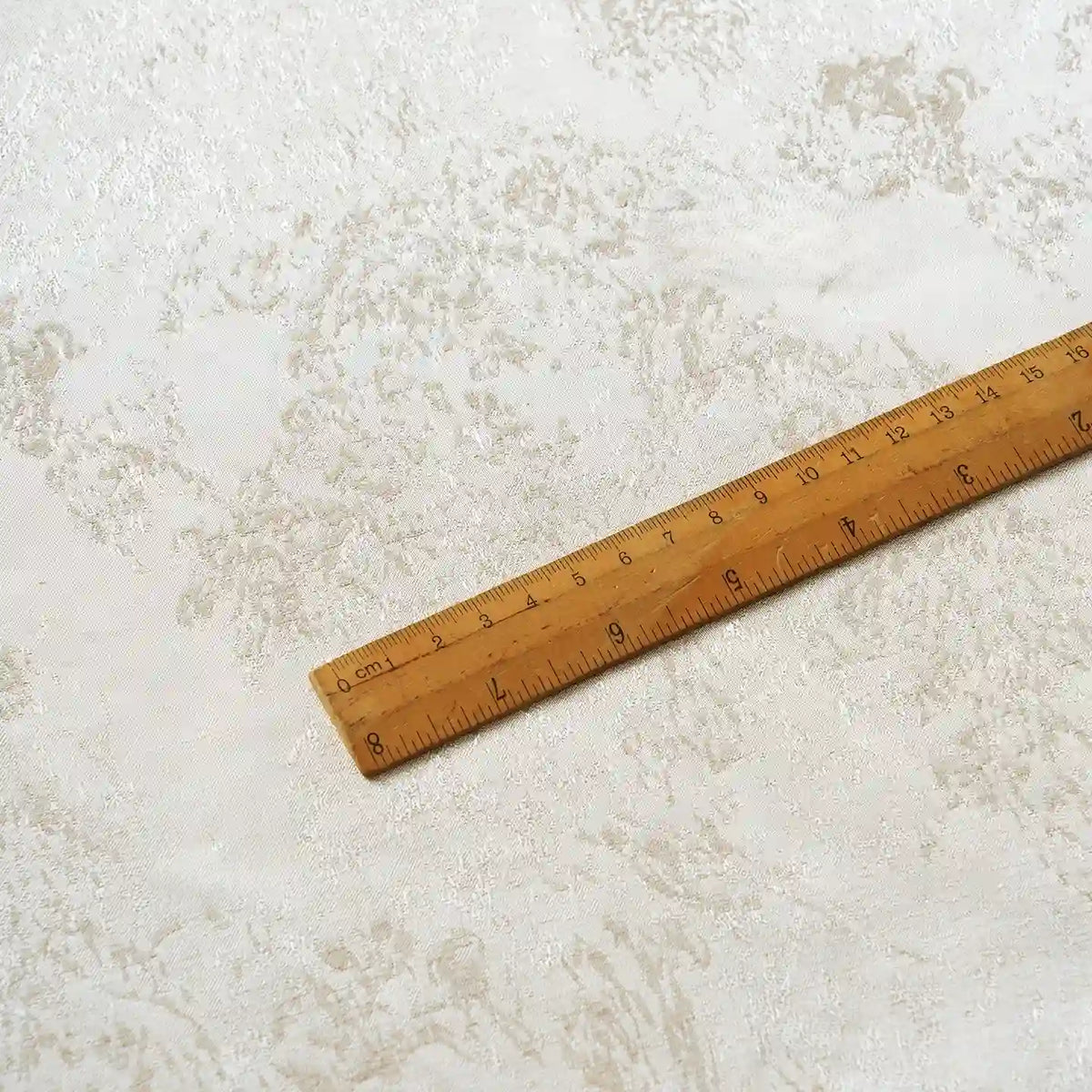 A wooden ruler marked in inches and centimeters lies diagonally across a light-colored, textured surface with subtle marbled patterns, reminiscent of the Linen Viscose Jacquard Satin Fabric by the yard from Longan Craft, featuring a subtle floral pattern and luxurious rayon satin material.