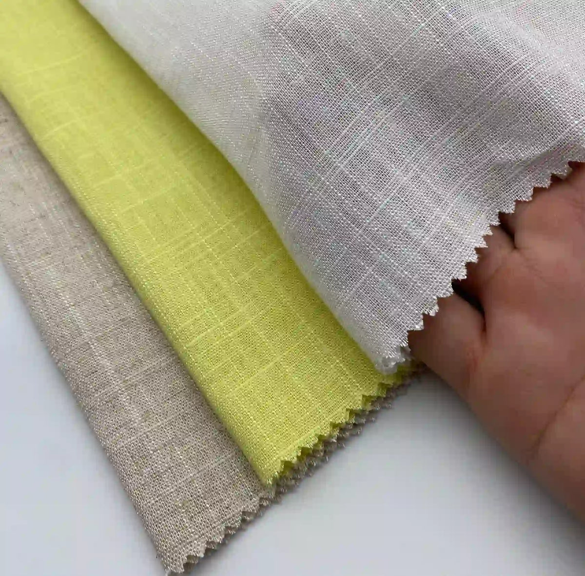 A hand displays three pieces of Longan Craft Linen and Viscose Fabric By The Yard in beige, yellow, and white—each with a textured, zigzag-cut edge. This breathable multicolor viscose cellulose fabric is ideal for summer projects.