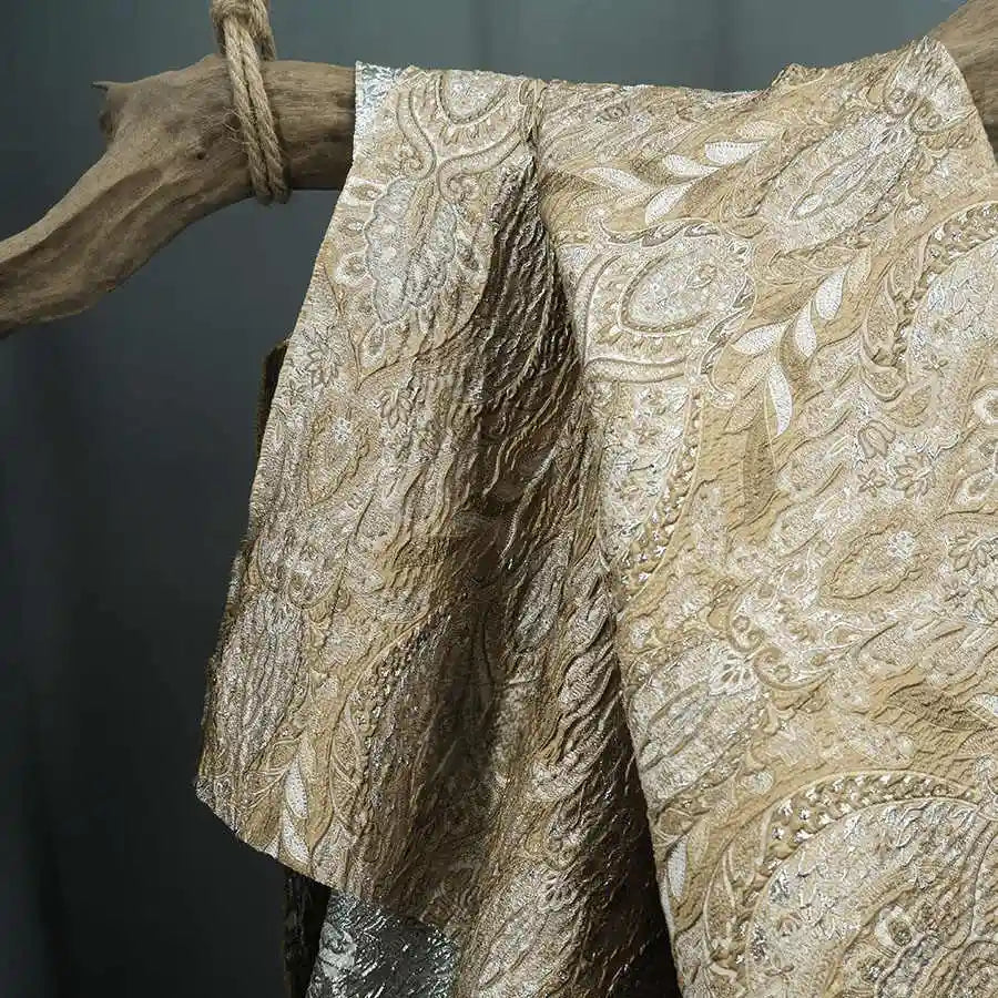 The Luxury Embossed Brocade Fabric by Longan Craft, a premium metallic jacquard with intricate paisley and floral designs in gold and silver, is draped elegantly over a rustic wooden branch tied with rope.